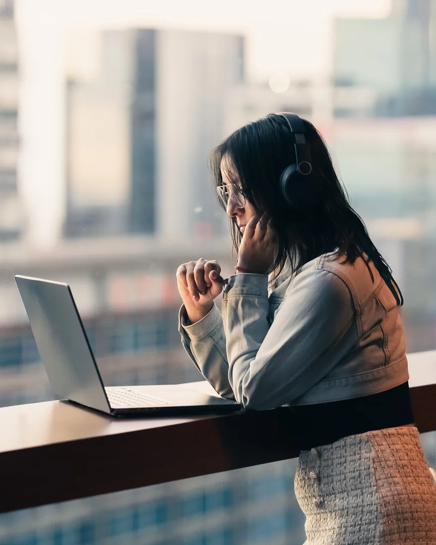 A founder at a high window counter above the business district, hand at her mouth, waiting on her laptop